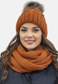 Cappello e sciarpa in arancio bruciato. Il cappello presenta una texture a costine e un pompon in pelliccia. La sciarpa ha un pattern a maglia spessa, che offre calore.