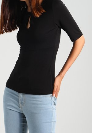 Femme portant un haut noir ajusté avec des manches au coude et un jean bleu clair, main reposant dans la poche arrière, sur un fond uni.