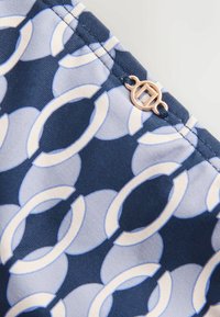 Tissu bleu et blanc avec motif de chaîne circulaire et une petite boucle en métal doré cousue près du bord cousu.