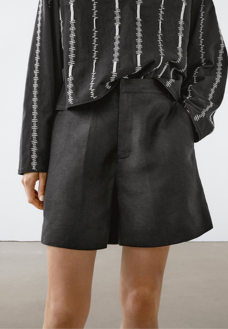 Short noir taille haute ajusté porté avec un haut noir à manches longues présentant des motifs de couture blanche verticale.