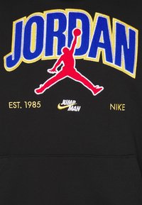 Svart hoodie med "JORDAN" i blå och gul brodyr, en röd Jumpman-logotyp samt "EST. 1985" och "NIKE" i gul brodyr.