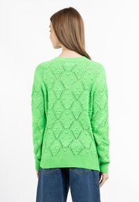Maglione verde lavorato a maglia con un motivo a diamante testurizzato, bordi a coste e orlo. La vista posteriore mostra una trama aperta e pompon decorativi.