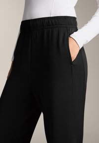 Čierne športové nohavice s elastickým pásom, bočnými vreckami a hladkou textúrou. Detailný záber zdôrazňujúci pás a detaily vreciek.