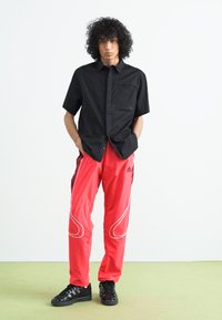 Sort kortærmet skjorte med knapper, røde sporty bukser med hvide detaljer og sorte sneakers. Modellen står foran en ensfarvet baggrund.