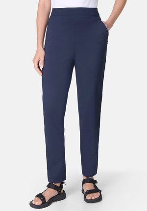 Vrouw draagt marineblauwe rechte broek met zijzakken en zwarte verstelbare sandalen, staand tegen een effen witte achtergrond.