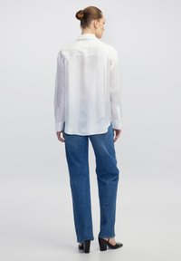 Biała satynowa bluzka z wiązaniem na plecach, długimi rękawami i mankietami na guziki, w zestawieniu z luźnymi niebieskimi dżinsami i czarnymi szpilkami.