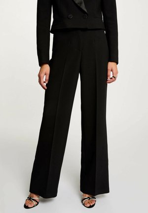 Donna che indossa pantaloni neri su misura a gamba larga, un blazer corto a doppiopetto nero e sandali neri con cinturini arricchiti da dettagli in argento.