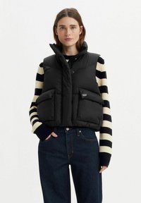 Schwarze Steppweste mit hohem Kragen, vorne mit Knopfverschluss und zwei großen Taschen. Über einem gestreiften Pullover getragen und kombiniert mit dunklen Jeans.