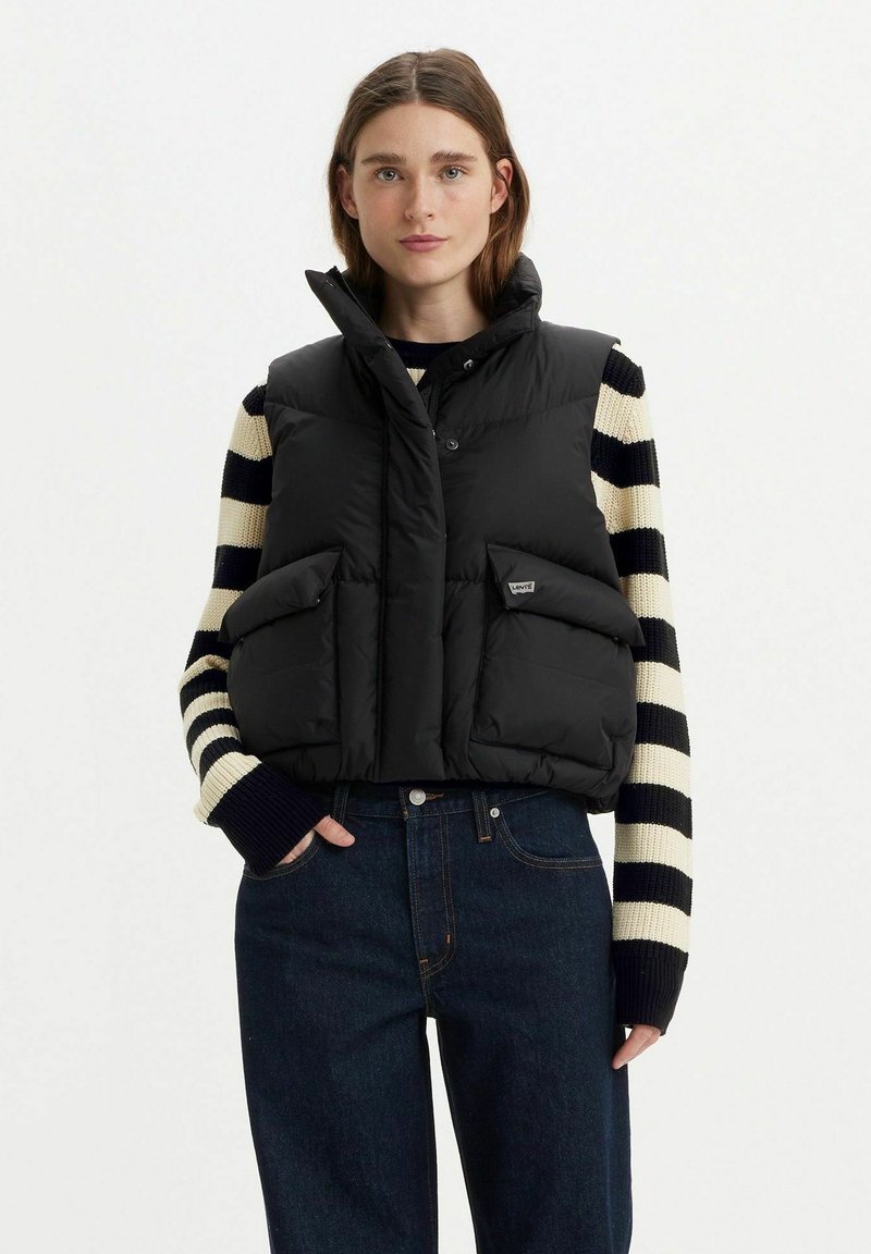 Gilet imbottito nero con colletto alto, chiusura frontale con bottoni e due grandi tasche. Indossato sopra un maglione a righe, abbinato a jeans scuri.