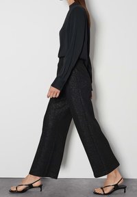 Kvinna i svart blus med långa ärmar och glittriga svarta vida byxor som går i svarta låga klacksandaler mot en vit vägg.