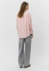 Ljusrosa långärmad skjorta med lös passform och rundad nederkant, tillsammans med lösa grå denimbyxor. Minimalistisk design och slät textur.