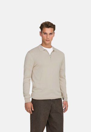 Jonge man die een beige poloshirt met lange mouwen en een voorrits draagt, met donkerbruine broek tegen een effen achtergrond.