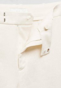 Pantalon en lin couleur crème avec une fermeture à bouton et deux fermoirs métalliques. Design épuré avec une texture lisse et des accents minimalistes en métal.