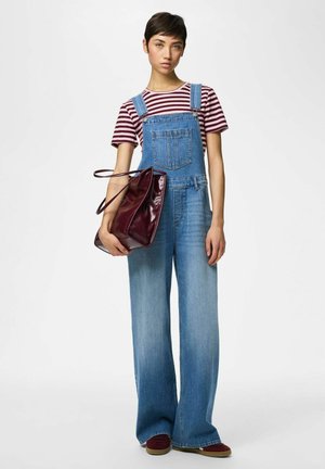 Fiatal személy csíkos pólót és bőszárú farmer napozót visel, nagy bordó táskát tart a kezében, fehér háttér előtt áll.