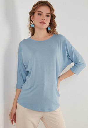 Blusa azzurro chiaro, comoda e ampia, con maniche a tre quarti, scollatura tonda, tessuto morbido e orlo curvo, abbinata a pantaloni beige.