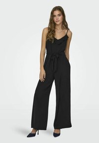 Zwart jumpsuit met een ontspannen pasvorm, ruches aan de halslijn, verstelbare spaghettibandjes, strikceintuur en wijde pijpen, gecombineerd met marineblauwe hakken.