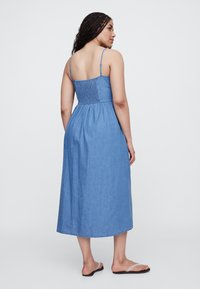 Plava midi haljina od trapera s tankim naramenicama, nabrano leđima i lepršavom suknjom. Ležerni stil sa mekanom teksturom, nošena s ravnim sandalama.