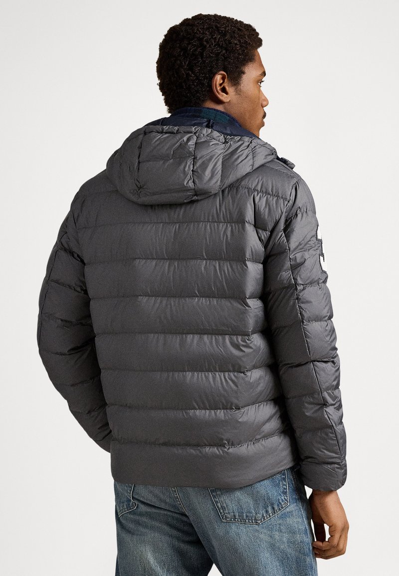 Doudoune grise avec capuche, dotée de sections matelassées et d'une texture lisse. Le design comprend une doublure intérieure contrastante et un patch logo latéral.