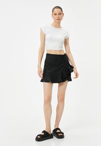 Witte korte t-shirt, strakke pasvorm. Zwarte gerimpelde minirok met zijverzameling. Zwarte plateau-sandalen met gekruiste bandjes.
