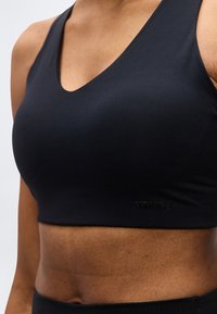 Κοντινό πλάνο ενός ατόμου που φοράει μαύρο αθλητικό σουτιέν με βαθύ ντεκολτέ και το όνομα της μάρκας "STRONGER" ανάγλυφο στο κάτω μέρος.