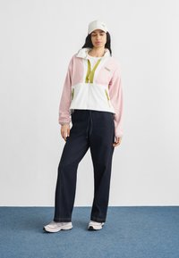 Σετ δύο τεμαχίων που περιλαμβάνει ένα ροζ και λευκό μπουφάν με φερμουάρ και πράσινες λεπτομέρειες, σε συνδυασμό με σκούρες μπλε παντελόνες με φαρδιά μπατζάκια και ανοιχτόχρωμα αθλητικά παπούτσια.