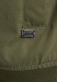 Patch din material verde cu margini cusute, având un eticheta metalică cu marca "Petrol". Textura este netedă cu finisaj mat.