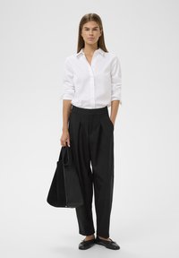 Valge nööpidega särk, mustad volditud püksid ja mustad korterkingad. Modell hoiab musta koti. Lihtne, struktureeritud disain puhtaid jooni.