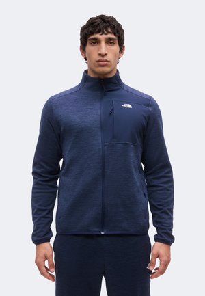 Mężczyzna stoi zwrócony przodem, ubrany w granatową kurtkę na zamek z logo The North Face na lewej kieszeni na piersi.