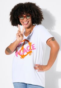 T-shirt blanc avec un graphique "No Peace" en rose et orange, manches courtes, coupe décontractée, porté avec un jean bleu. Cheveux bouclés et lunettes orange.