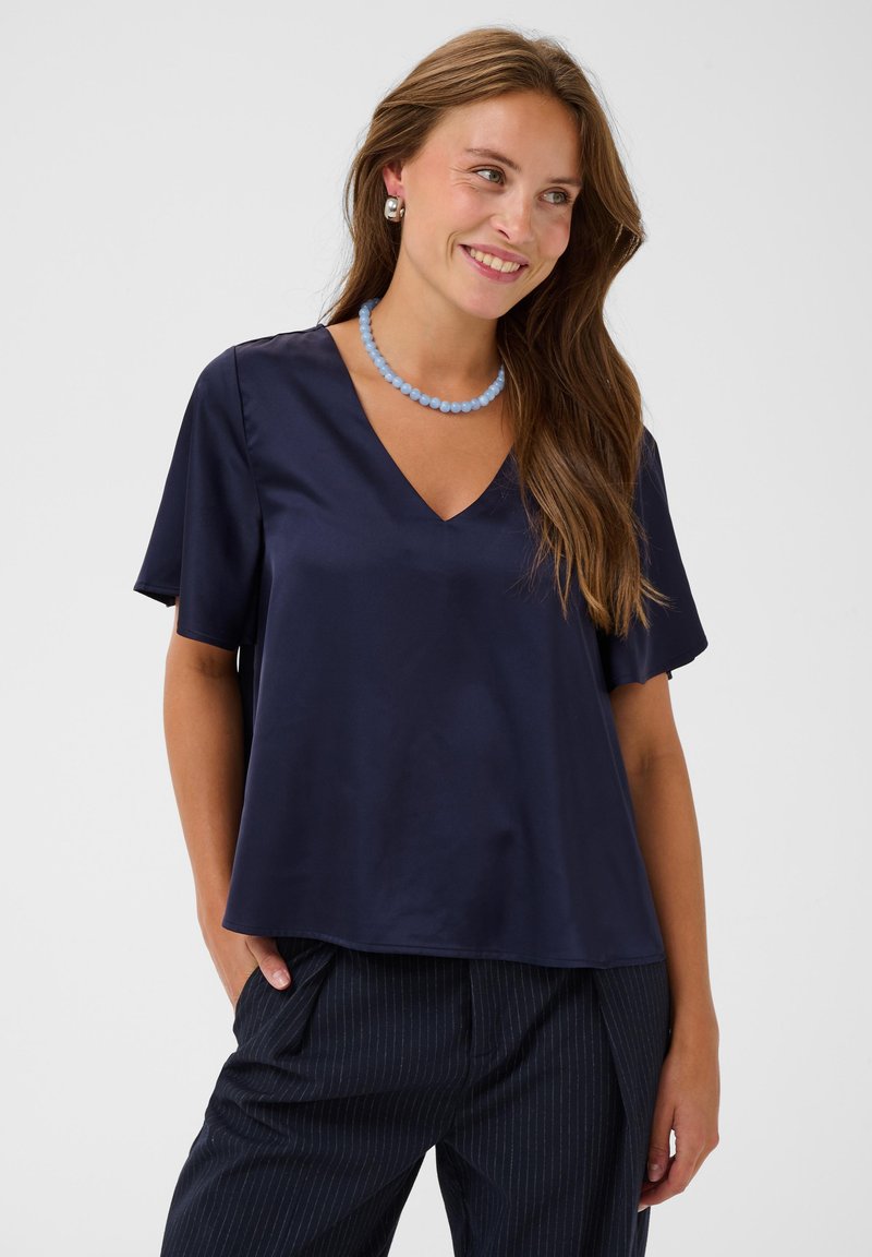 Femme souriante portant un chemisier ample bleu marine à col en V, pantalon à fines rayures, collier de perles bleu, boucles d'oreilles argentées, cheveux longs et bruns, mains dans les poches.