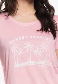 Kvinde iført en lyserød skjorte med hvide palmer og teksten "SUNSET ESCAPE" og "ROXY" trykt på fronten.
