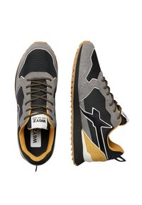 Un paio di sneakers nere e grigie con accenti giallo senape, suole testurizzate, lacci neri e gialli, e un logo bianco sui lati e sulla linguetta.
