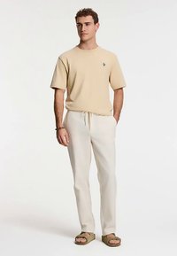 Beige Kurzarm-T-Shirt mit kleinem blauem Logo, kombiniert mit cremefarbenen Jogginghosen und grünen Sandalen. Einfacher, lässiger Schnitt.