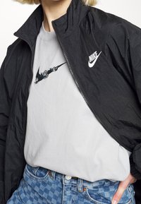 Person som bär en svart Nike zip-up jacka över en grå T-shirt med en blommig Nike swoosh-design och blå rutig jeans.