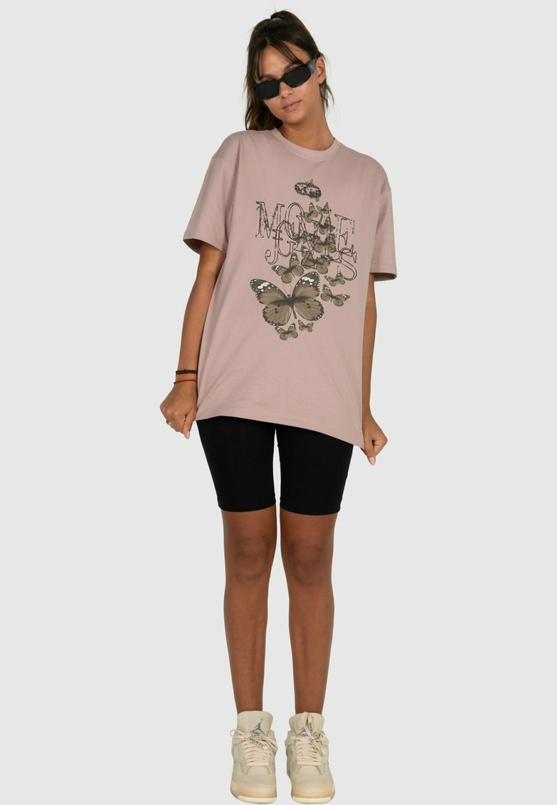 Růžové oversized tričko s motivem motýla a textem, spárované s černými upnutými šortkami a krémovými teniskami. Krátké rukávy a volný střih.