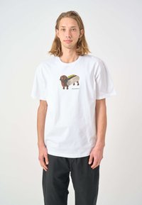 White cotton t-shirt featuring a colorful illustration of a dachshund with a hot dog. Short sleeves and relaxed fit. Black pants worn below.