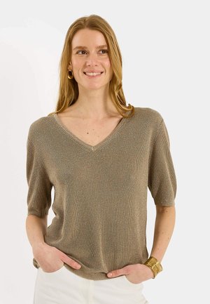 Femme aux longs cheveux blonds portant un haut métallique à manches courtes en V et un pantalon blanc, souriante, les mains dans les poches, sur fond blanc.