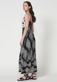 Robe maxi noire avec motif paisley blanc, présentant un corsage en crochet, des bretelles ajustables et une jupe fluide. Assortie à des sandales noires.