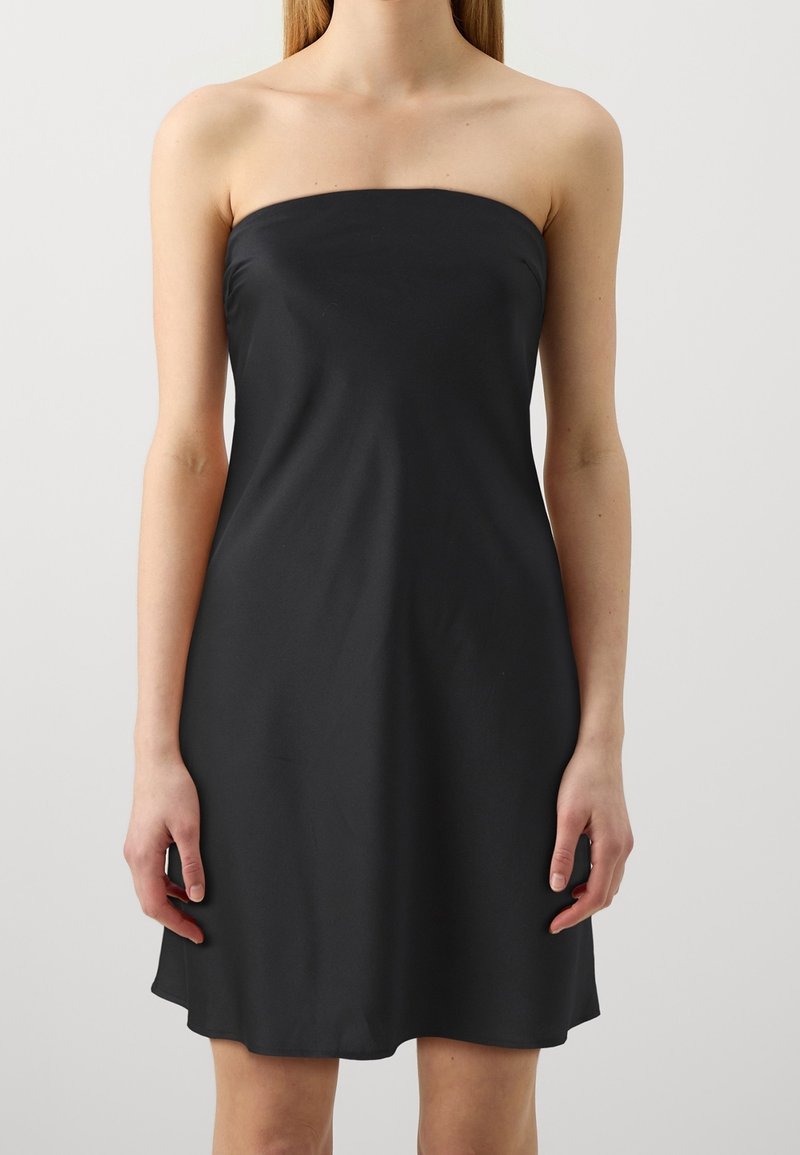 Robe noire sans bretelles portée par une personne debout, les bras relâchés le long du corps, montrée de la mi-cuisse aux épaules sur un fond uni.