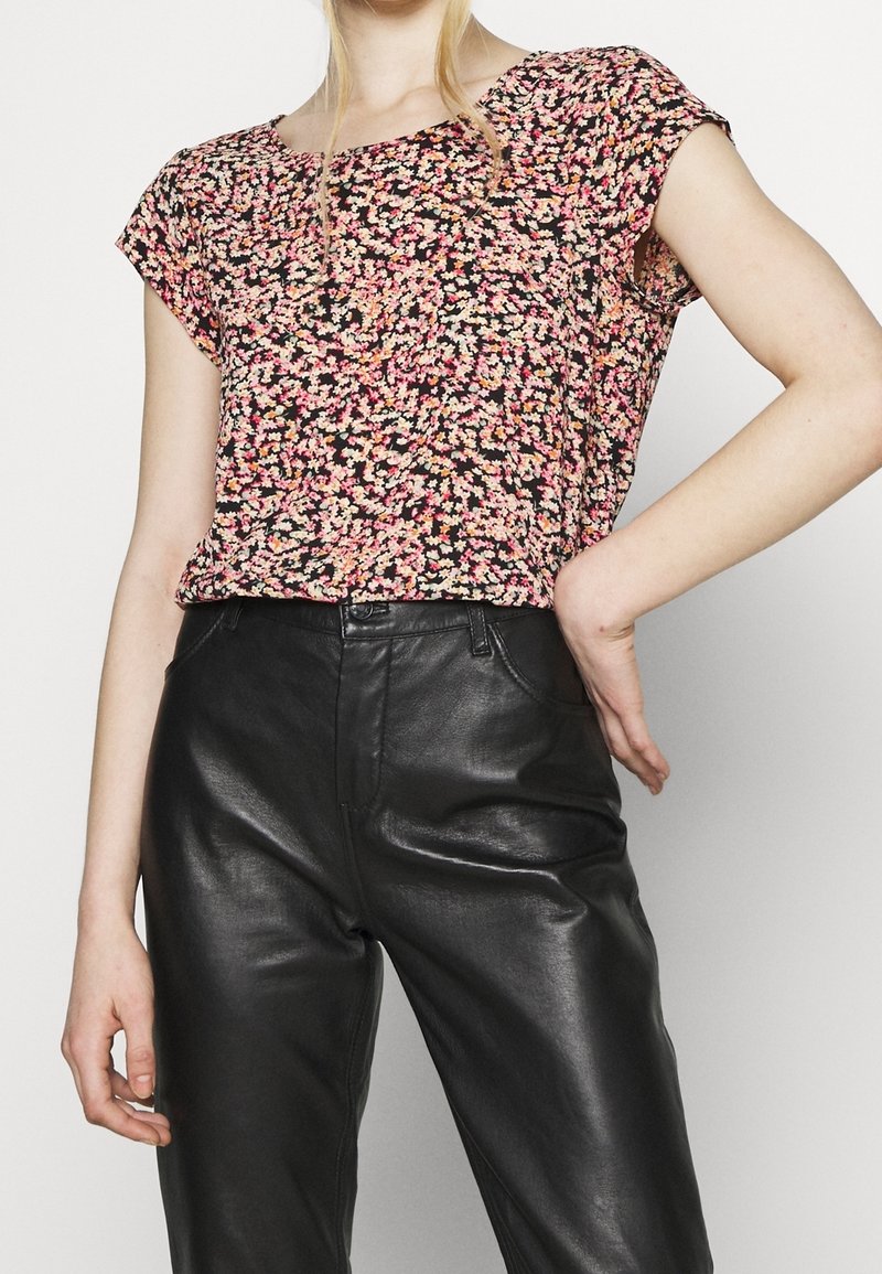 Femme portant un haut à manches courtes à fleurs roses et noires, rentré dans un pantalon en cuir noir taille haute, debout avec une main sur la hanche.
