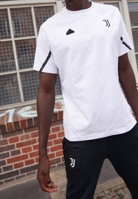 Biała koszulka sportowa z czarnymi akcentami, z logo Adidas na piersi oraz emblematem Juventusu. W połączeniu z czarnymi spodniami z logo.