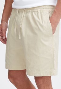 Beige shorts made of lightweight fabric with a textured finish. Features an elastic waistband with drawstring and side pockets.