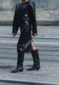 Giacca cropped in pelle marrone con bottoni, abbinata a una gonna a tubino in pelle marrone con uno spacco laterale e stivali neri al ginocchio con tacco.
