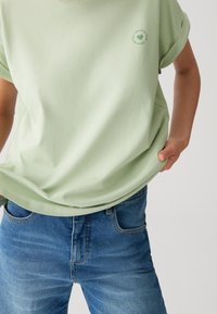 T-shirt in cotone verde chiaro con maniche arrotolate e un dettaglio rotondo del logo sul davanti, abbinato a jeans blu in denim.