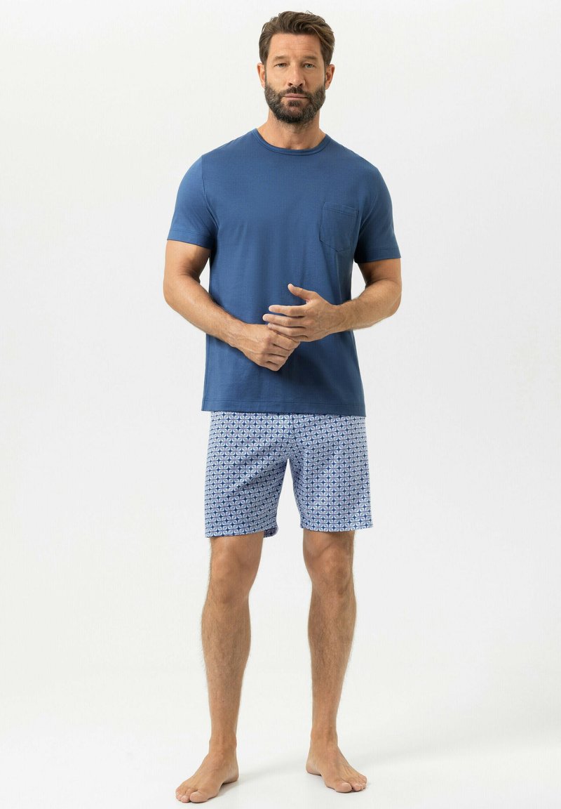 Mann steht barfuß da, trägt ein schlichtes blaues kurzärmeliges Hemd und gemusterte blaue und weiße Shorts, die Hände vor der Taille verschränkt.