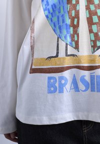 Λευκό πουκάμισο με μακριά μανίκια, με πολύχρωμο σχέδιο πουλιού που στέκεται πάνω σε κίτρινη μπάρα και το κείμενο "BRASIL" από κάτω, φορεμένο με σκούρο τζιν.