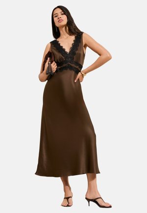 Kobieta w brązowej sukience midi z satyny z czarną koronkową lamówką, trzymająca dopasowaną kopertówkę, nosząca brązowe sandały na obcasie i złotą bransoletkę.
