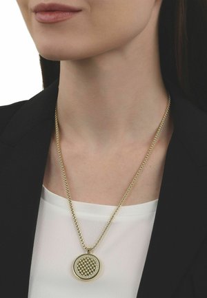 Frau trägt eine goldene Halskette mit rundem Anhänger, der ein Wabengittermuster und kleine Kristalle aufweist, gekleidet in einem weißen Oberteil und schwarzem Blazer.