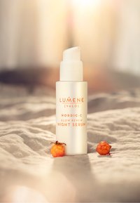 Lumene Nordic-C Glow Renew Night Serum i en frostad glasflaska med en vit pump, accentuerad med orange text. Två orange bär i närheten.