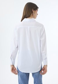 Weißes Langarmhemd mit Kragen, Rückenfalten-Detail und offenen Ärmelschlitzen. Kombiniert mit hellblauen Jeans.